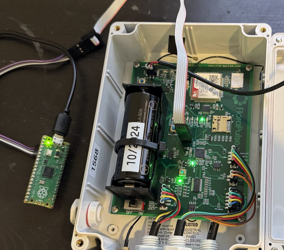 The complete setup: the updater board on the left with its USB cable and green light on, its ribbon cable running into the opened FloraPulse logger, which sits open with its battery and circuit board visible